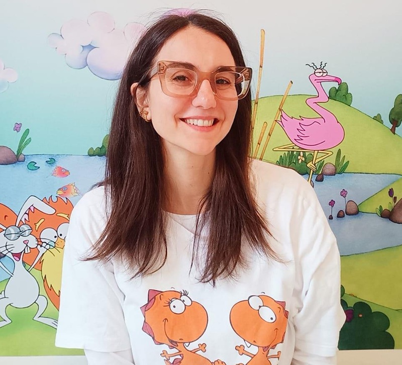 Valentina Stolfa sorridente con occhiali rosa e maglietta bianca con draghetti arancioni davanti a un murale con fenicottero